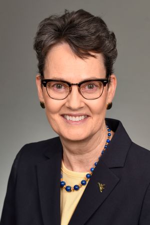 Female with short dark hair wears glasses, circle earrings, a blue blazer with a WVU pin.