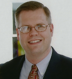 Headshot of a man with short hair, glasses, and suit smiling
